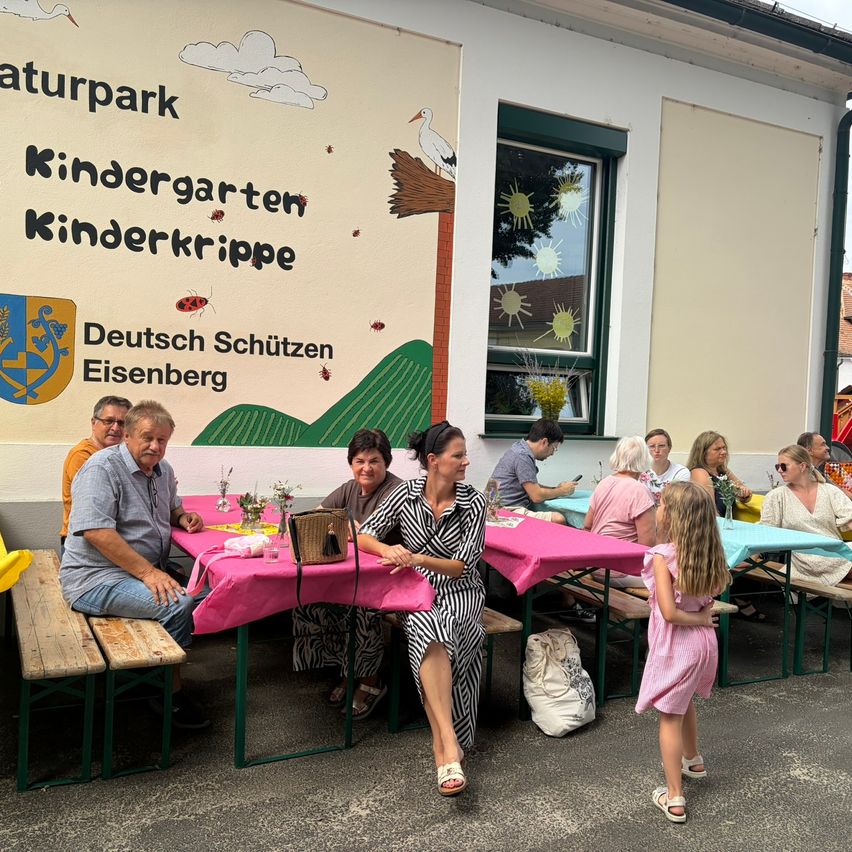 A group of people sit at a picnic table outside a building with a mural and the text Kindergarten Kinderkrippe. They are eating and drinking. A little girl stands in front of them.