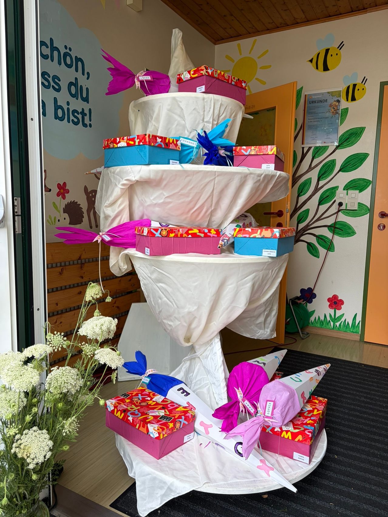 A multi-layered cake-like display of wrapped gifts with colorful ribbons is set on a table covered in white cloth. There are flowers on the left and a wall with drawings on the right.