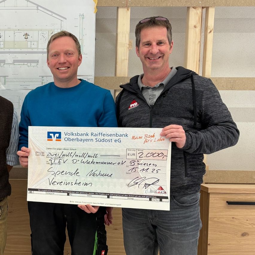 Drei Männer stehen lächelnd vor einer Holzwand. Ein Mann hält einen großen Scheck mit dem Betrag von 20.000 Euro. Hinter ihnen befindet sich eine Tafel mit Architekturzeichnungen.