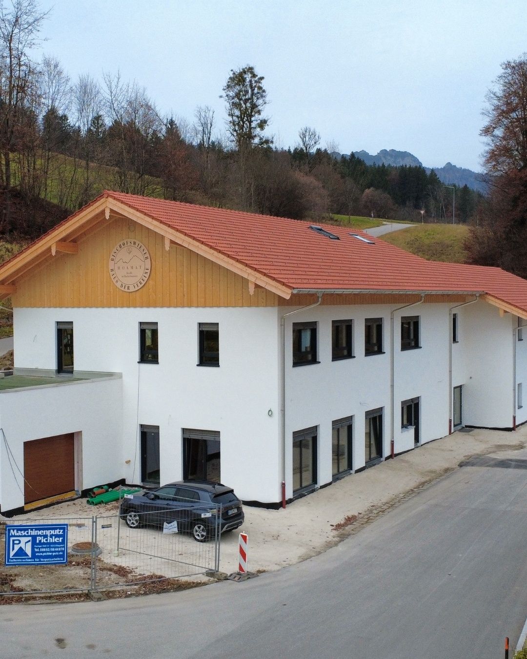 Ein modernes weißes Gebäude mit rotem Dach wird gebaut. Ein schwarzes Auto ist davor geparkt. Das Gebäude hat ein Logo über dem Eingang.