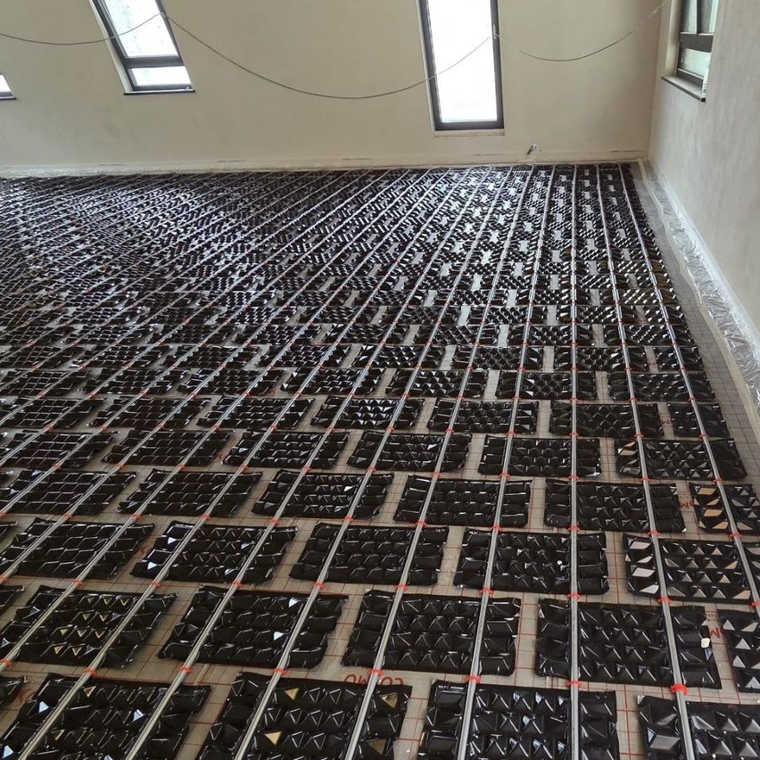 Ein mit schwarzen Fliesen bedeckter Boden ist von oben zu sehen. Die Fliesen sind in einem Gittermuster angeordnet, wobei einige Fliesen ein geometrisches Design aufweisen. Der Boden ist leer, und es sind keine Menschen oder Gegenstände zu sehen.