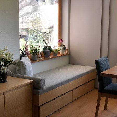 Eine gemütliche Ecke mit einer Bank unter einem Fenster, einem hölzernen Tisch mit zwei Stühlen und einer Vase mit weißen Blumen auf einem Schrank. Auf der Fensterbank stehen Topfpflanzen.