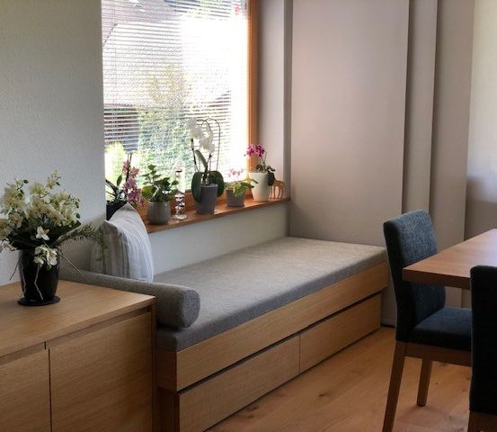 Eine gemütliche Ecke mit einer Bank unter einem Fenster, einem hölzernen Tisch mit zwei Stühlen und einer Vase mit weißen Blumen auf einem Schrank. Auf der Fensterbank stehen Topfpflanzen.