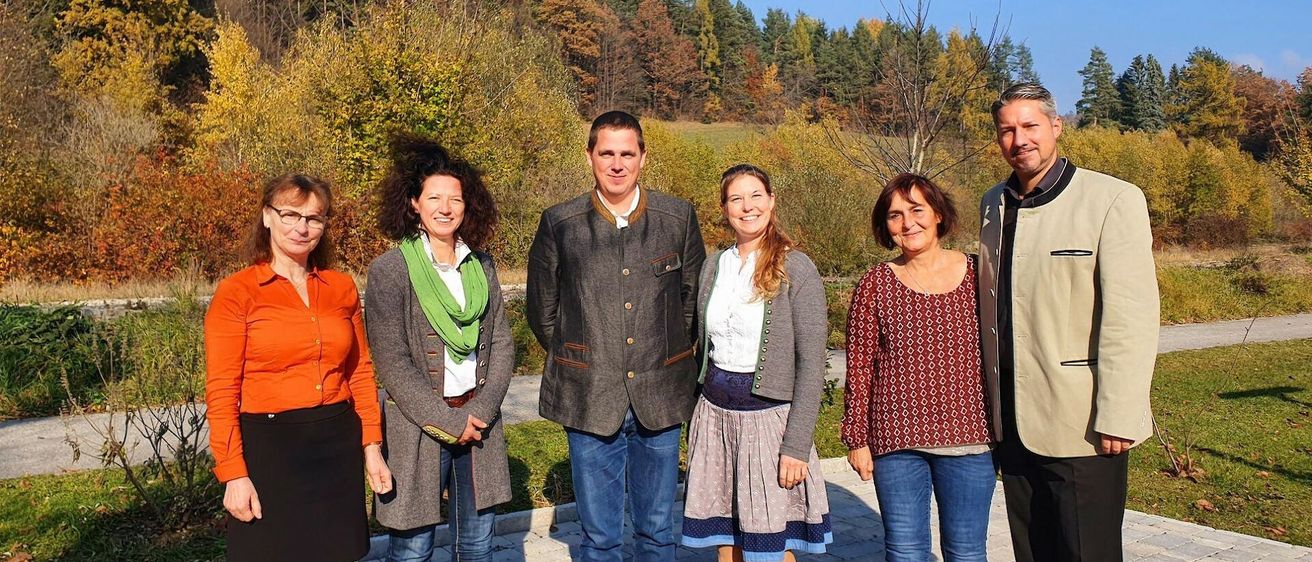 Eine Gruppe von sechs Personen steht auf einem Ziegelweg in einem Park mit herbstlichen Bäumen im Hintergrund.