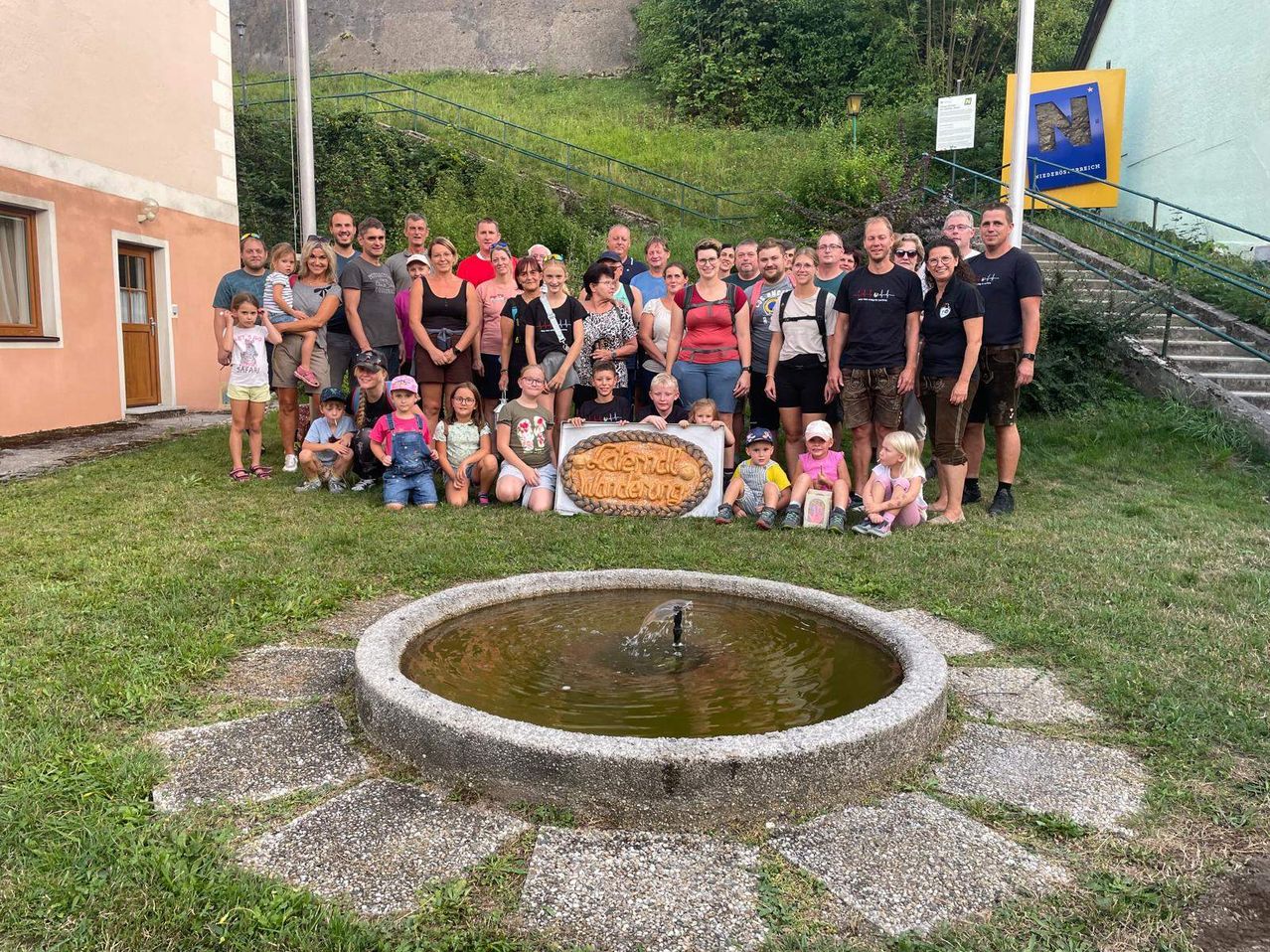 Eine Gruppe von Menschen, einschließlich Erwachsener und Kinder, posiert für ein Foto vor einem Brunnen und einem Schild.