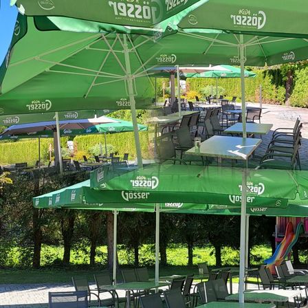 Ein Außenbereich mit mehreren grünen Sonnenschirmen, Tischen und Stühlen. Die Sonnenschirme haben den Text '19220' darauf. Im Hintergrund befinden sich Bäume und eine Rutsche.