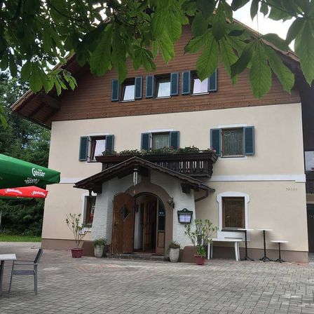 Ein zweistöckiges Gebäude mit braunen Holzverkleidungen und blauen Fensterläden ist zu sehen. Es hat einen Balkon mit Blumentöpfen, einem grünen Schirm und Topfpflanzen. Draußen gibt es Tische und Stühle.