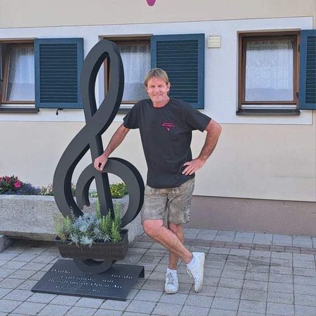Ein Mann steht neben einer großen musikalischen Noten-Skulptur vor einem Gebäude mit einem Hirschlogo und dem Wort 'Wirtshaus'.