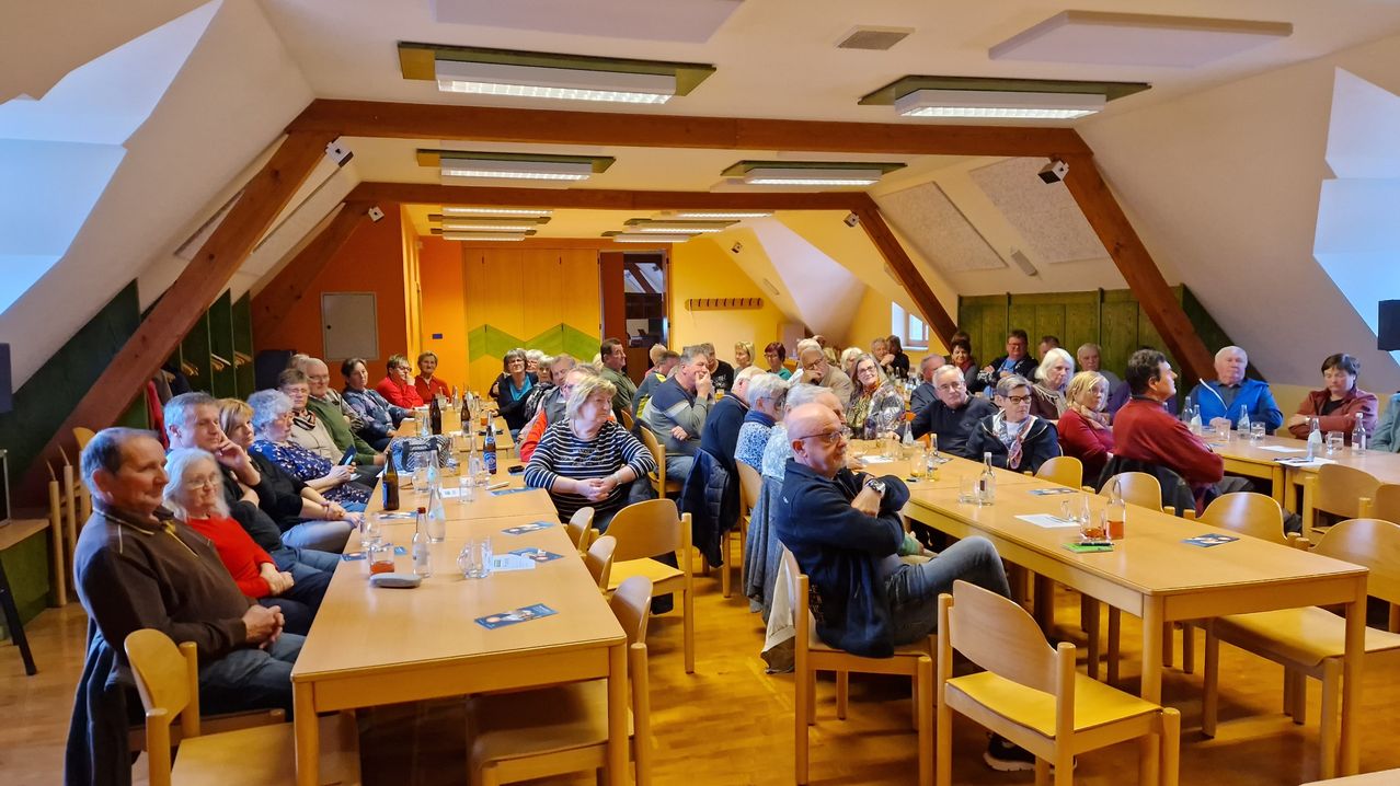 Eine große Gruppe von Menschen sitzt an Tischen in einem Konferenzraum. Die Tische sind mit Flaschen, Gläsern und Papieren gefüllt. Der Raum hat Holzböden, eine gelbe Wand und eine Decke mit Lampen.