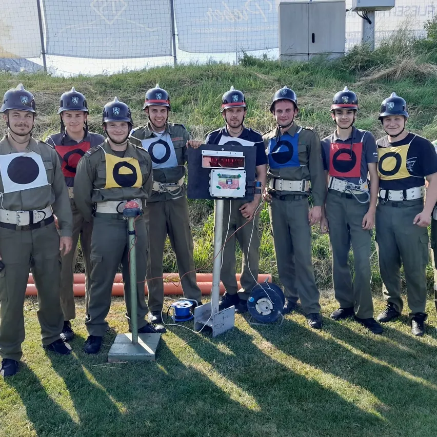 Eine Gruppe von Soldaten in Uniformen mit verschiedenfarbigen Zielen auf der Brust, stehen auf einem Rasenfeld. Ein digitales Scoreboard steht vor ihnen.