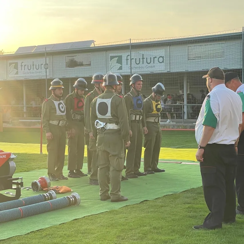 Eine Gruppe von Personen in Uniformen steht auf einem grünen Feld. Sie tragen Helme und einige haben Gürtel. In der Nähe beobachten zwei Männer in Mützen und einer in einem grünen Hemd. Ausrüstung und ein Banner mit der Aufschrift 'frutura' sind vorhanden.