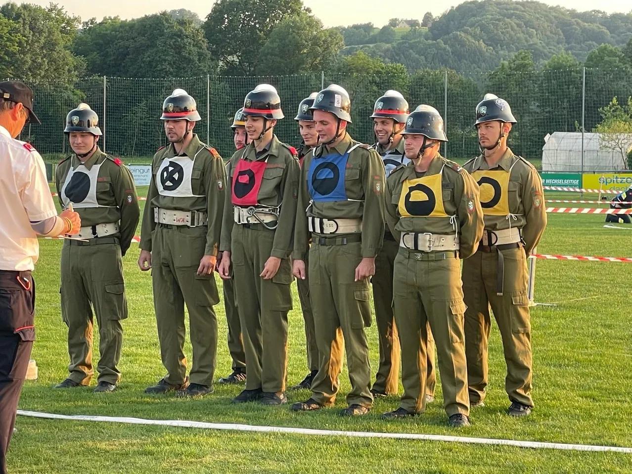 Eine Gruppe von Männern in Militäruniformen steht auf einem Grasfeld, wahrscheinlich bei einer militärischen Trainingsveranstaltung. Sie tragen Helme und Gürtel mit verschiedenen Symbolen. Im Hintergrund befinden sich ein Zaun und Bäume.