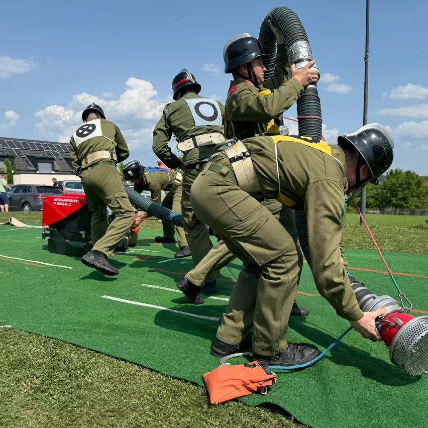 Eine Gruppe von Menschen in Uniformen arbeitet auf einem grünen Feld mit einer Feuerwehrspritze. Sie tragen Helme, schwarze Schuhe und Gürtel. Sie machen eine Art Training.