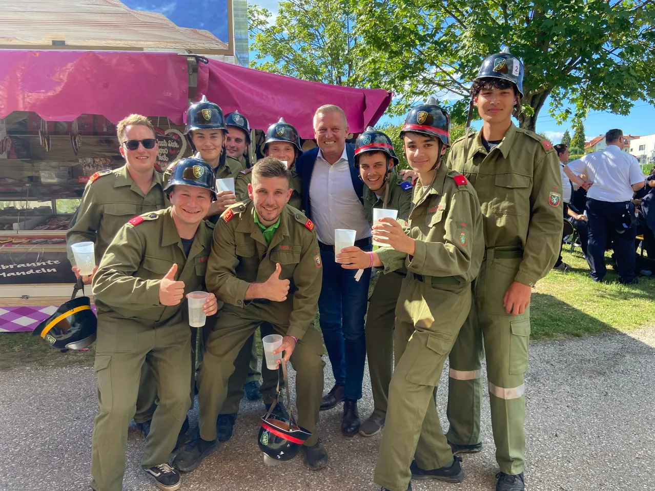 Eine Gruppe von Menschen in Militäruniformen posiert für ein Foto mit einem Mann in einem weißen Hemd, unter einem rosa Zelt, mit Bäumen und einem Gebäude im Hintergrund.