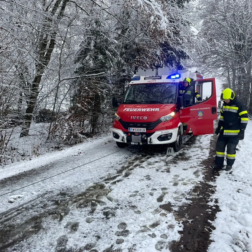 Ein roter Feuerwehrwagen mit offener Tür steht auf einer verschneiten Straße. Ein Feuerwehrmann steht daneben, beide tragen gelbe Helme.
