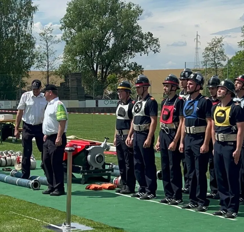 Eine Gruppe Feuerwehrleute in Uniform steht auf einem Feld mit Bäumen und einem Zaun im Hintergrund. Zwei Männer in weißen Uniformen stehen in der Nähe.