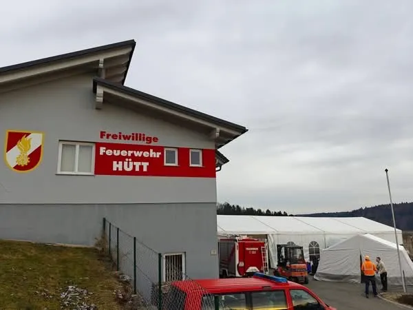 Ein graues Gebäude mit der Aufschrift Freiwillige Feuerwehr Hutt. Im Hintergrund steht ein Zelt, an dem Leute stehen. Vor dem Gebäude ist ein rotes Fahrzeug geparkt.