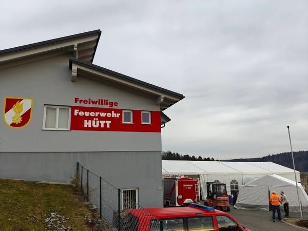 A gray building with the words Freiwillige Feuerwehr Hutt on it. There is a tent in the back with people standing near it. There is a red vehicle parked in front of the building.