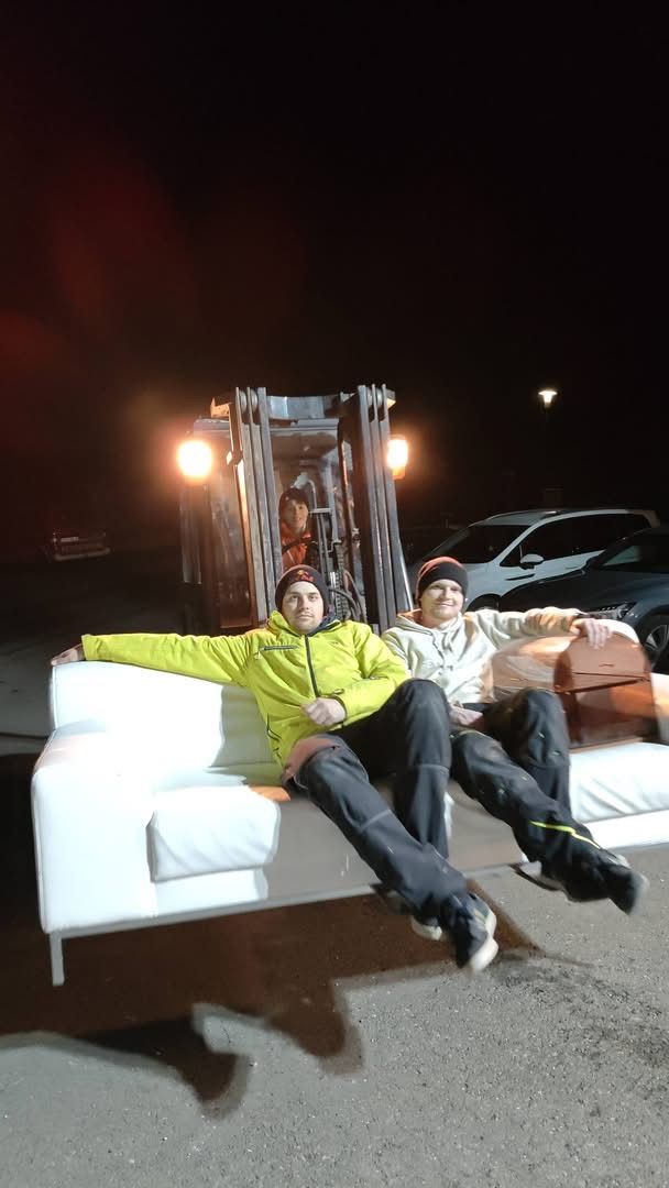 Two men sit on a white couch at night. A forklift is behind them with a person sitting inside. There are cars parked behind the forklift.