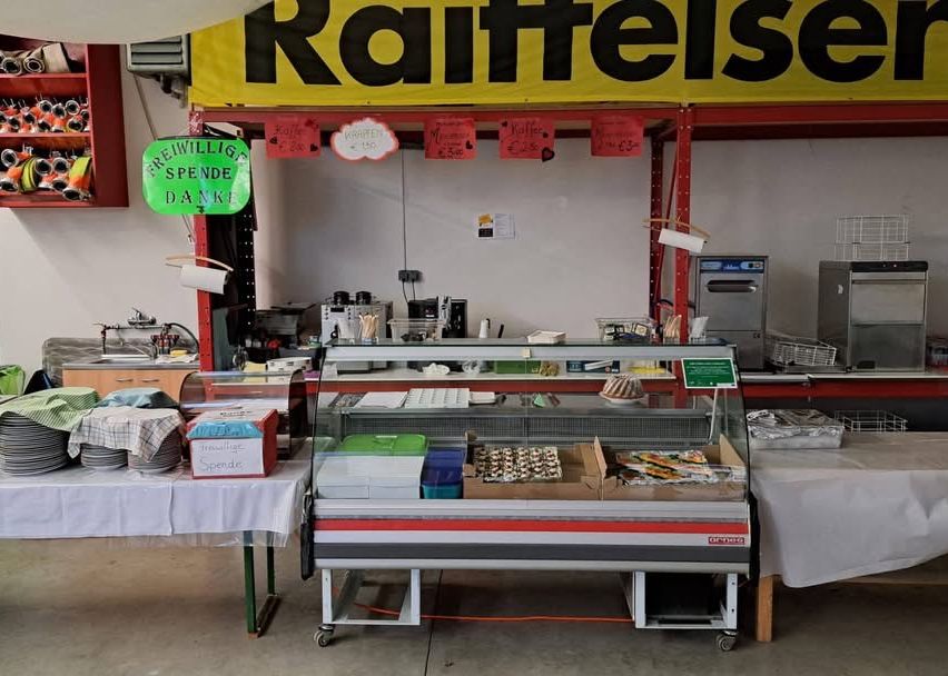 A display case with pastries sits in front of a counter with a sign that says 'Freiwillige Spenden Danke'. The counter has various items on it. A shelf is mounted on the wall. Another table is covered with a white cloth and has plates on it.