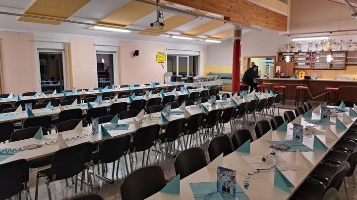 An empty banquet hall with long tables, each set with a blue napkin and a menu card. The room has a counter with stools, a bar area, and a large window. A person is standing near the counter.