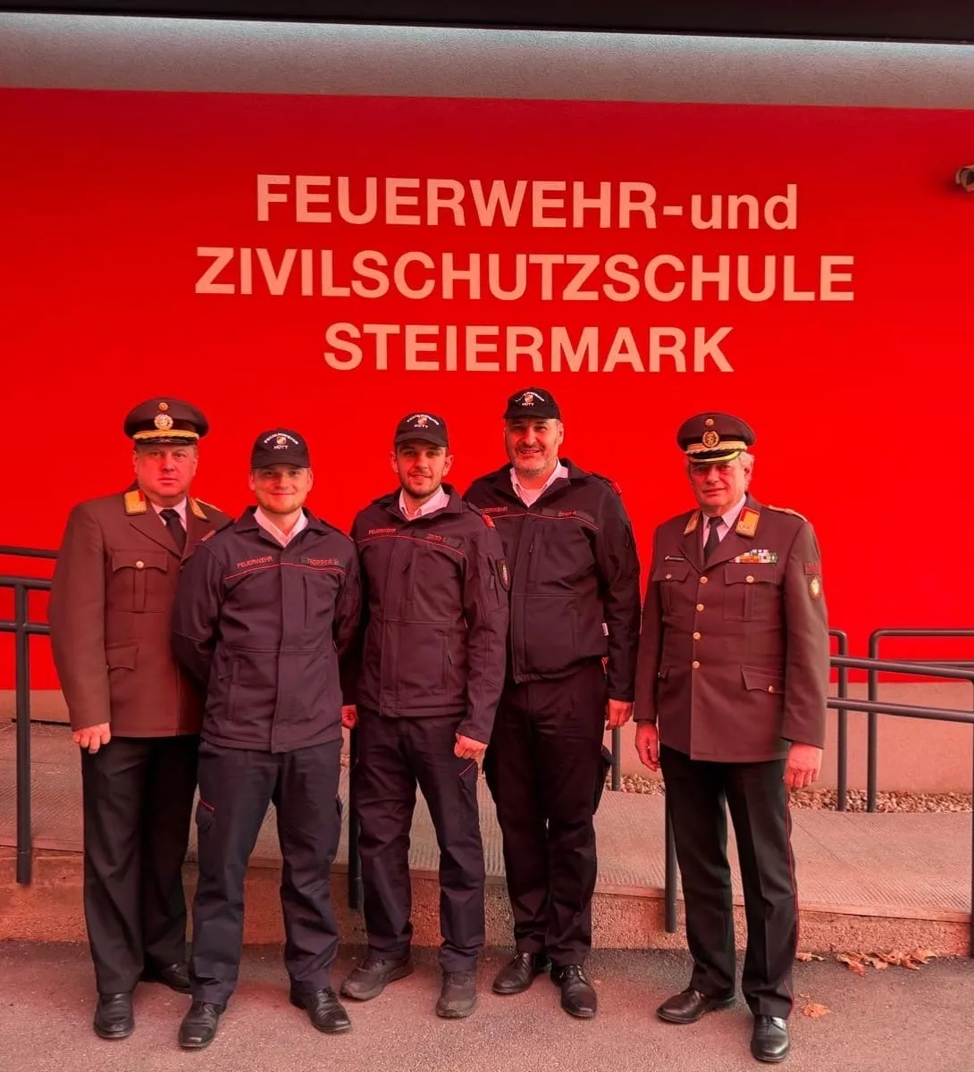Fünf Männer in Uniform stehen vor einer roten Wand. Es steht Feuerwehr-und Zivilschutzschule Steiermark. Sie lächeln und posieren für ein Foto.