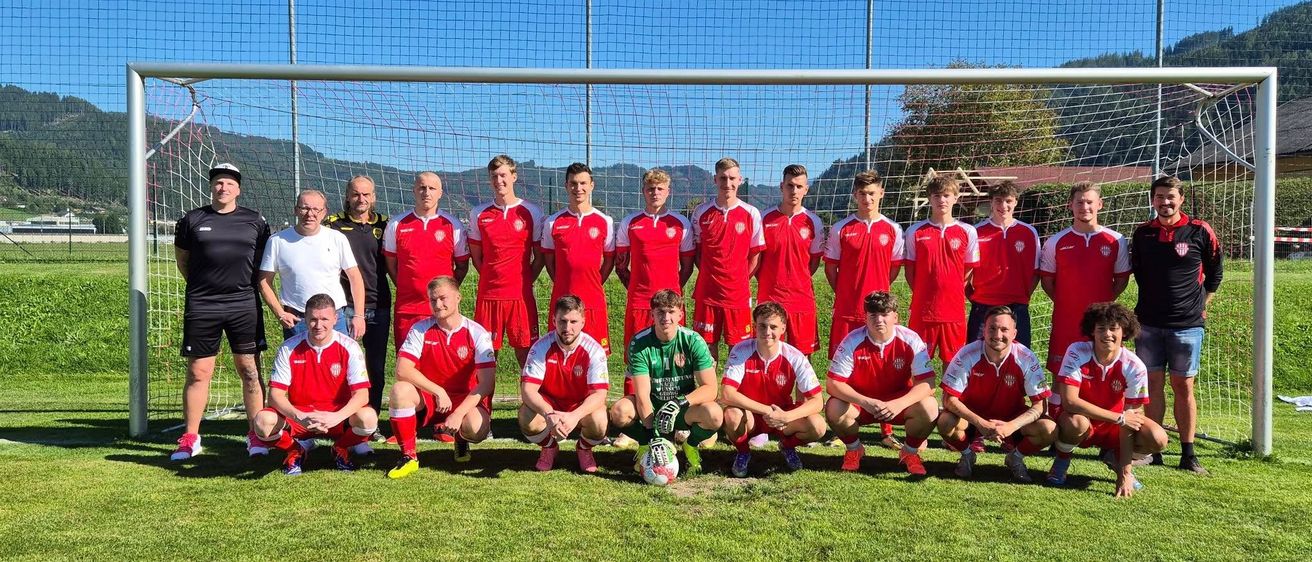 Eine Fußballmannschaft in roten und weißen Trikots posiert vor einem Tor. Der Torwart in Grün steht neben dem Ball. Dahinter eine Metallgitter und ein Rasenfeld mit Bergen in der Ferne.