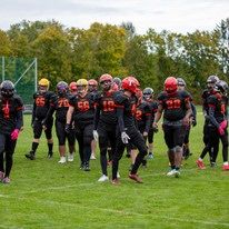 Eine Gruppe von Footballspielern in schwarzen Trikots, Helmen und Handschuhen läuft über ein Feld. Die Spieler sind in einer Reihe aufgestellt, möglicherweise in Vorbereitung auf ein Spiel.
