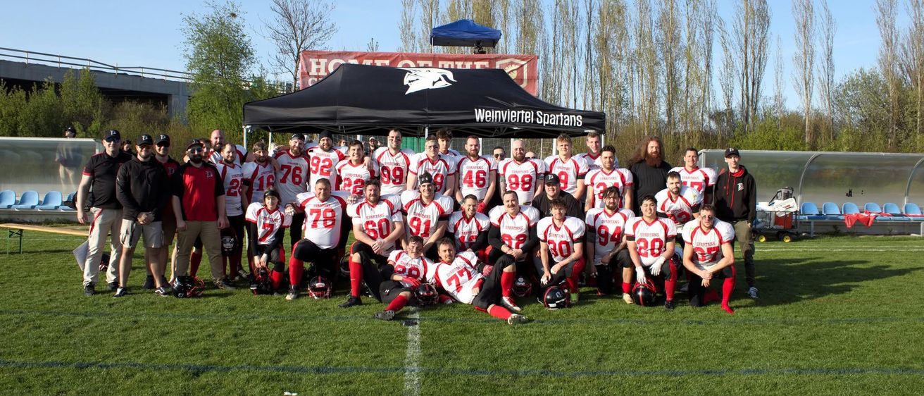Das Weinviertel Spartans Football-Team posiert für ein Foto auf einem Feld, mit einem schwarzen Zelt hinter ihnen. Sie tragen weiße Trikots mit Nummern und rote Hosen.