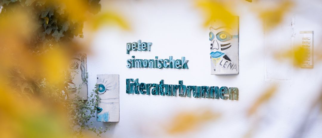 Eine weiße Wand mit einem braunen Dach zeigt Schilder mit 'peter simonischek' und 'literaturbrunnen'. Ein quadratisches Schild mit einem Gesicht ist darüber. Unscharfe Blätter und Pflanzen sind im Vordergrund.