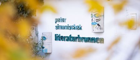 Eine weiße Wand mit einem braunen Dach zeigt Schilder mit 'peter simonischek' und 'literaturbrunnen'. Ein quadratisches Schild mit einem Gesicht ist darüber. Unscharfe Blätter und Pflanzen sind im Vordergrund.