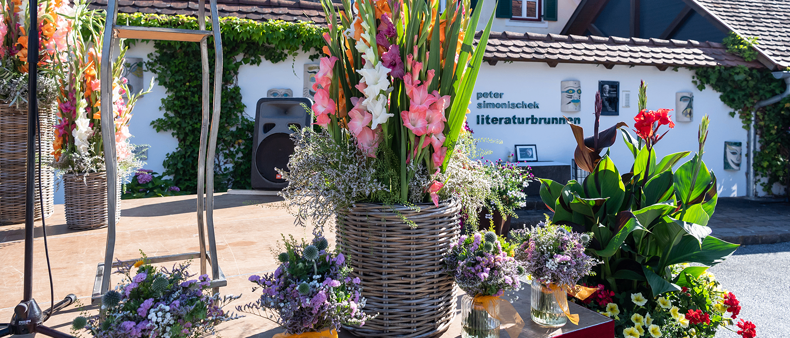 Eine Bühne mit einem großen Korb aus Weide mit bunten Blumen und drei kleinen Vasen mit Blumen. Hinter der Bühne steht ein Haus mit dem Namen Peter Simonischek Literaturbrunnen.