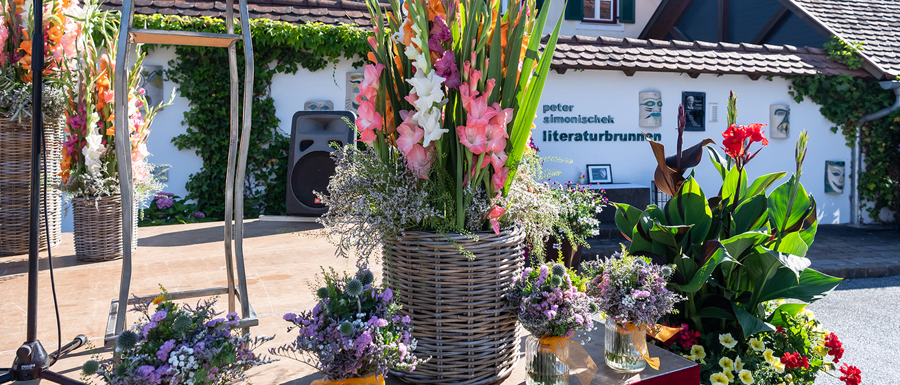 Eine Bühne mit einem großen Korb aus Weide mit bunten Blumen und drei kleinen Vasen mit Blumen. Hinter der Bühne steht ein Haus mit dem Namen Peter Simonischek Literaturbrunnen.