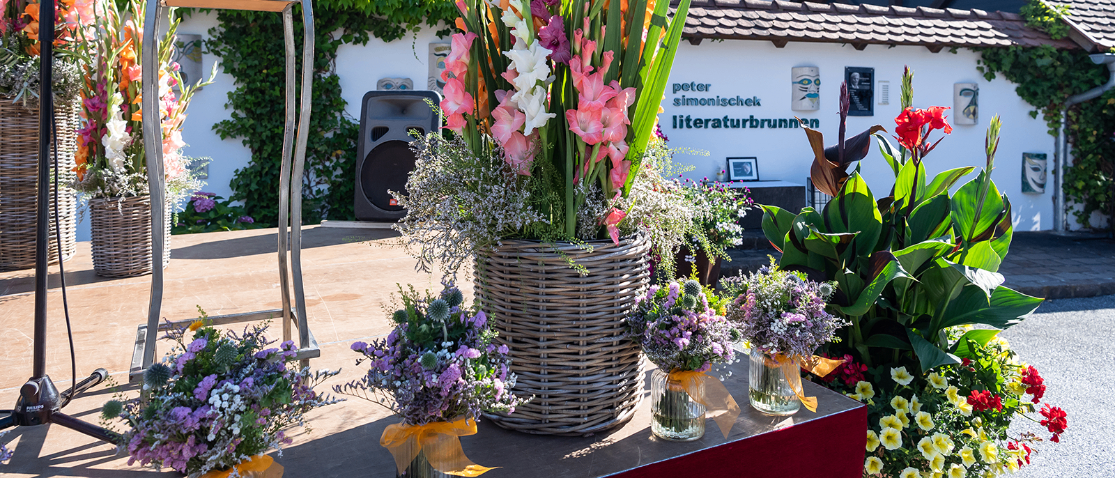 Eine Bühne mit einem großen Korb aus Weide mit bunten Blumen und drei kleinen Vasen mit Blumen. Hinter der Bühne steht ein Haus mit dem Namen Peter Simonischek Literaturbrunnen.