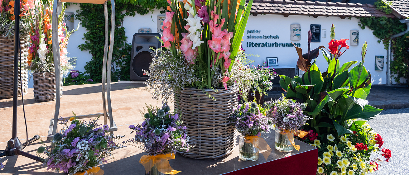 Eine Bühne mit einem großen Korb aus Weide mit bunten Blumen und drei kleinen Vasen mit Blumen. Hinter der Bühne steht ein Haus mit dem Namen Peter Simonischek Literaturbrunnen.