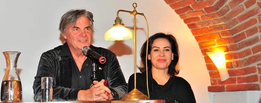 Ein Mann und eine Frau sitzen an einem Tisch mit Mikrofon und Lampe. Sie lächeln beide und schauen zur Kamera. Der Hintergrund besteht aus einer Backsteinmauer.