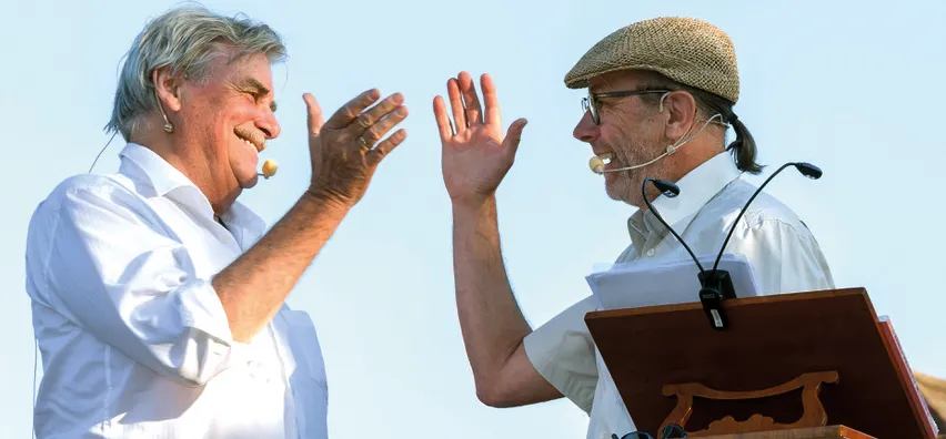 Zwei Männer lächeln und gestikulieren, während sie Kopfhörer tragen und vor einem Podium stehen. Der Mann auf der linken Seite hat ein Mikrofon im Mund.