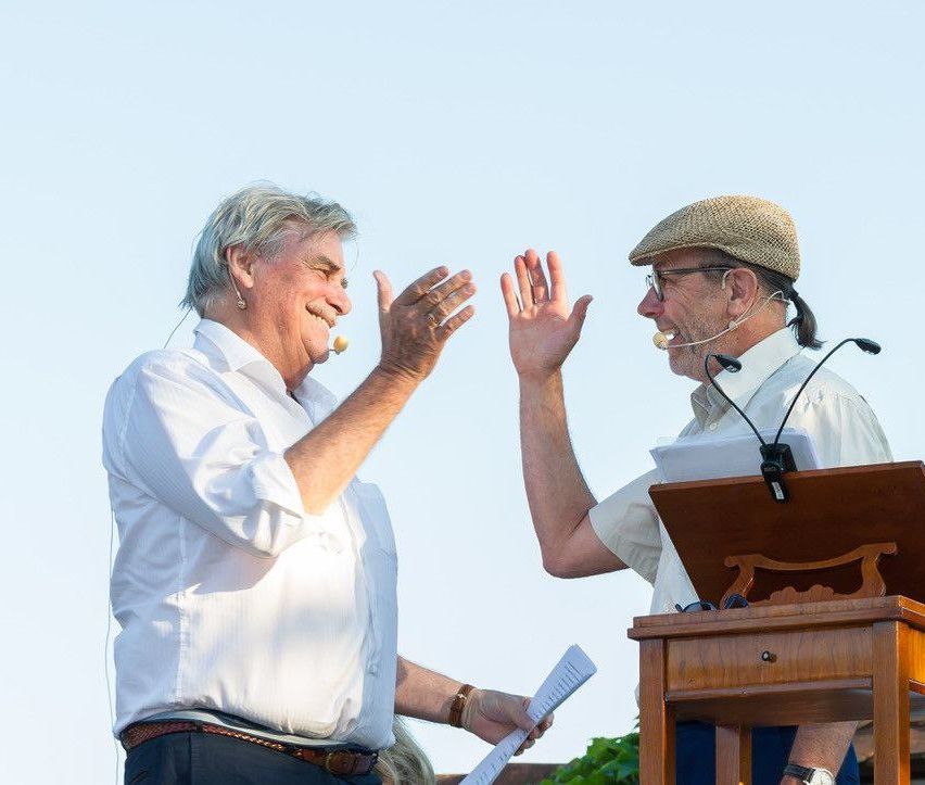 Zwei Männer stehen an einem Rednerpult, einer hält ein Papier in der Hand, beide tragen Hüte und Mikrofone, lächeln und winken, als würden sie eine Rede oder Präsentation beenden.