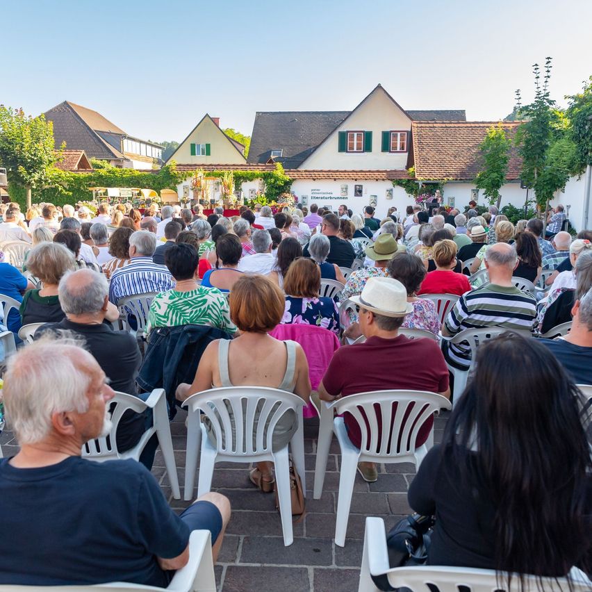 Eine Menschenmenge sitzt auf Stühlen im Freien. Einige tragen Hüte und Schuhe. Der Bereich ist von Häusern mit grünen Fenstern umgeben.