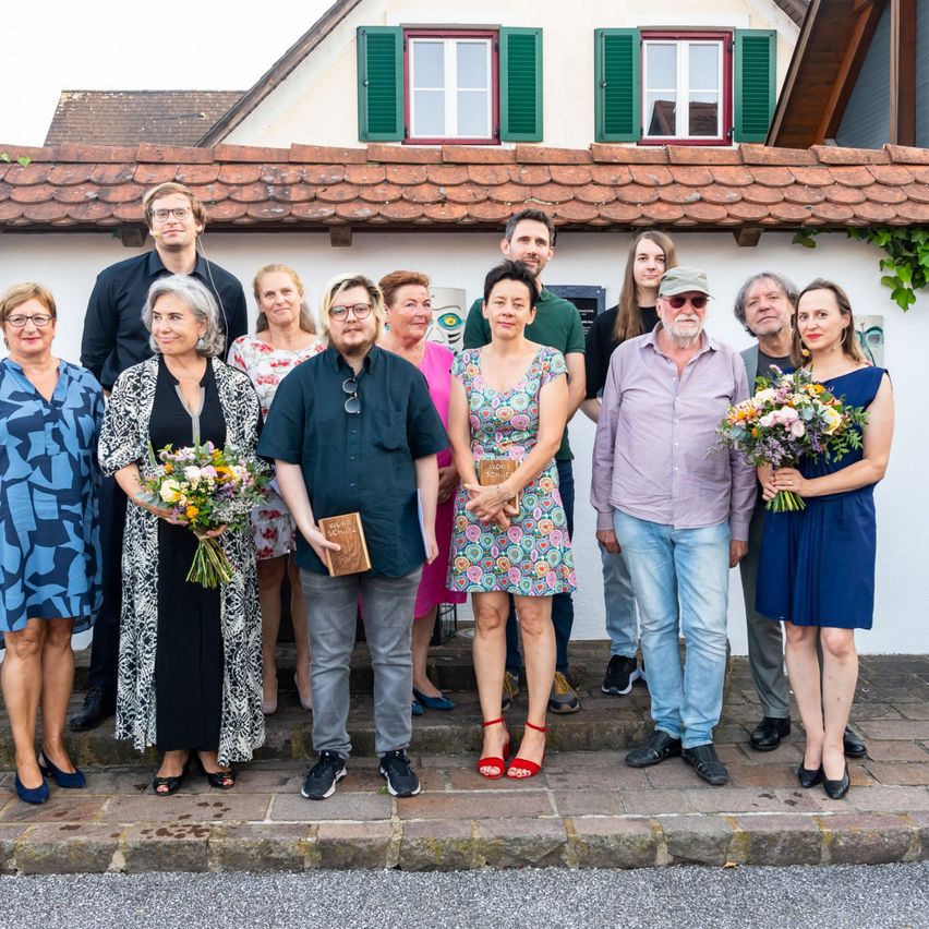 Eine Gruppe von Menschen in Freizeitkleidung steht vor einem Haus mit Ziegeldach und grünen Fensterläden. Sie halten Blumen und ein Buch.