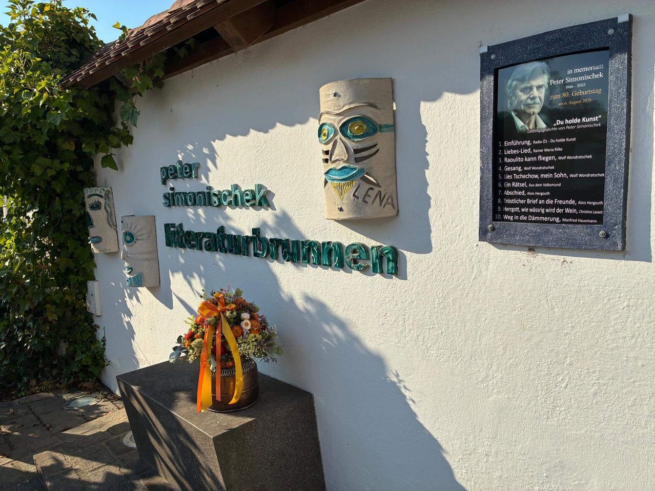 Eine weiße Wand mit grünem Text, der 'peter simonischek' und 'Literaturbrunnen' liest. Eine hölzerne Maske mit dem Namen 'LENA' ist an der Wand befestigt. Eine Plakette mit einem Bild eines Mannes und einigem Text ist ebenfalls angebracht. Ein Blumenstrauß ist auf einem Betonsockel vor der Wand platziert.