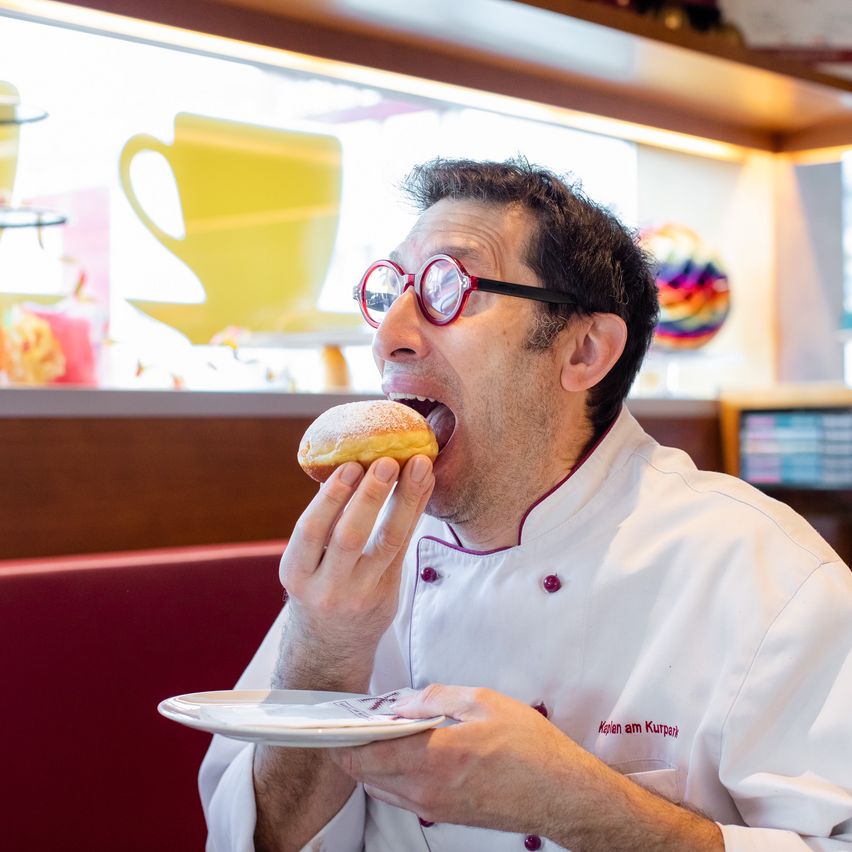 Ein Koch in einer weißen Uniform und mit einer Brille isst einen Donut von einem Teller. Im Hintergrund gibt es eine Ausstellung von bunten Gebäckstücken und einer gelben Tasse.