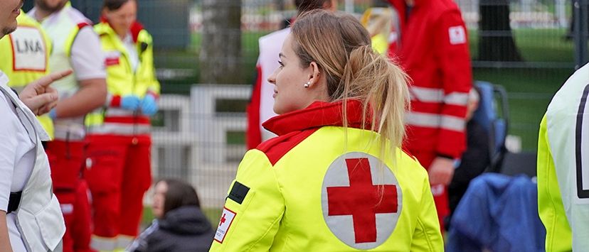Eine Frau in einer gelben und roten Jacke mit rotem Kreuzemblem steht im Freien mit mehreren anderen Personen in Sicherheitsausrüstung, möglicherweise bei einer medizinischen Veranstaltung.