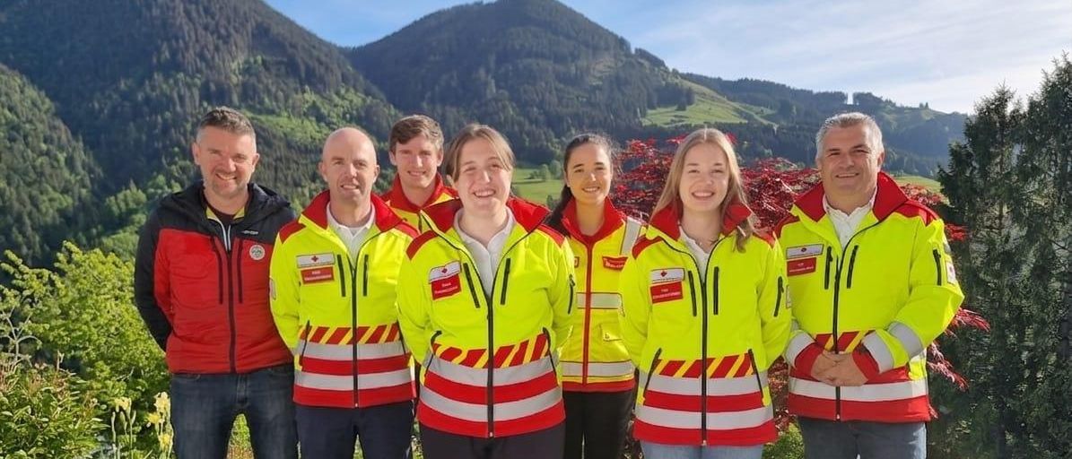 Eine Gruppe von sieben Personen in leuchtend gelben Westen mit roten und weißen Streifen steht gemeinsam auf einem Rasenfeld und lächelt für ein Foto vor Bergen.