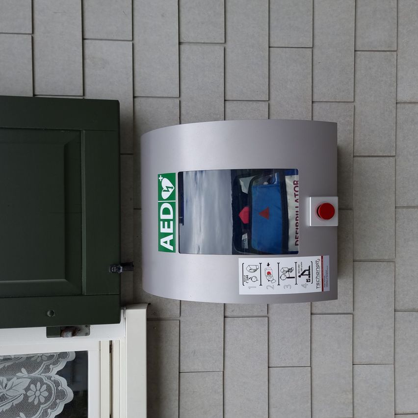 Ein AED-Gerät an einer Wand montiert mit einer grünen Tür links und einem Fenster mit Spitzenvorhang rechts.