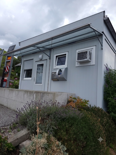 Ein graues Gebäude mit einem Glasdach und einer Klimaanlage an der Wand. Es hat eine Tür und zwei Fenster, eines mit einem Rollladen. Vor dem Gebäude stehen Pflanzen.