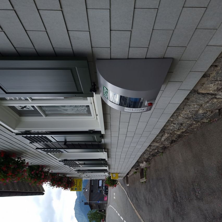 Ein Gebäude mit grauen Fliesen und einer grauen Metallbox über einer Tür. Die Box hat einen roten Knopf und ein grünes Ausfahrtsschild.