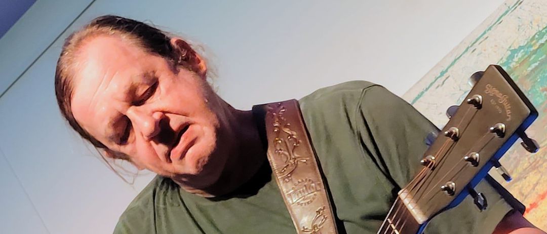 Ein Mann in einem grünen T-Shirt spielt eine Gitarre mit einem braunen Gurt. Die Augen des Mannes sind geschlossen. Ein Mikrofon befindet sich links vom Mann.