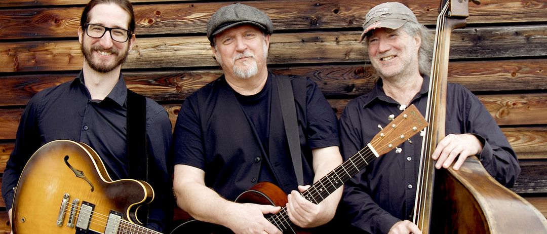 Drei Männer in lässiger Kleidung stehen vor einer Holzwand, lächelnd und Gitarren haltend. Der Mann links trägt eine Brille, die Mitte hält einen Bass und der Rechte trägt eine Mütze.