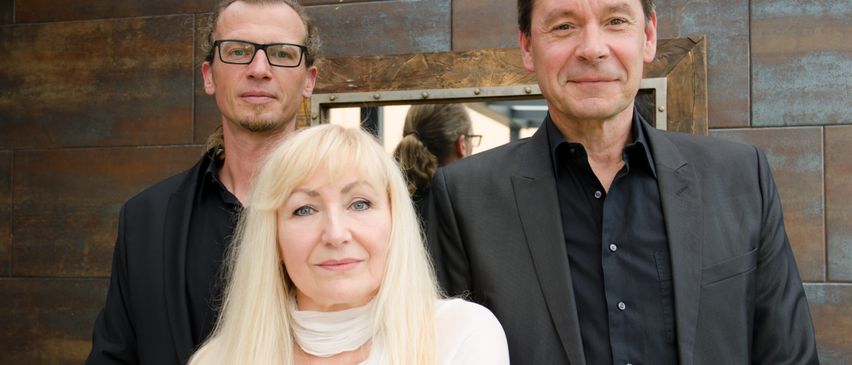 Drei Personen stehen vor einer Metallwand, eine Frau in Weiß und zwei Männer in schwarzen Anzügen. Der Mann links trägt eine Brille.